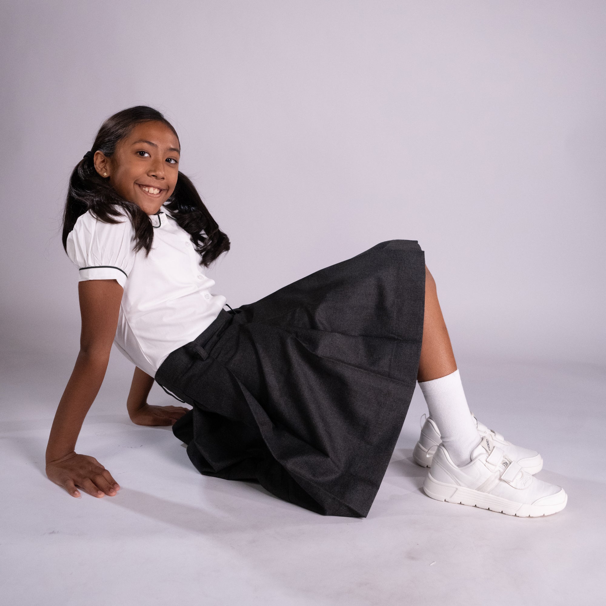 Young girl sitting on a white surface wearing a white shirt, black skirt, and white sneakers.